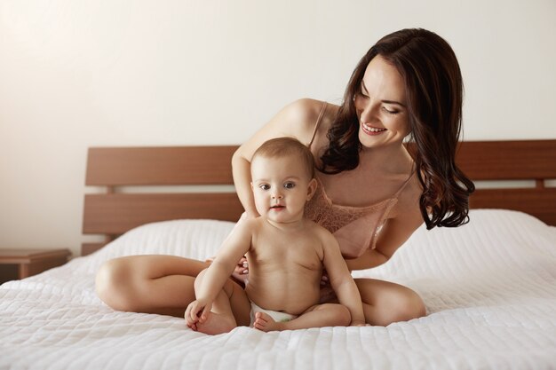 Neugeborenes Baby sitzt zu Hause mit ihrer Mutter, die spielt und lächelt. Mutter genießt ihre Mutterschaft.