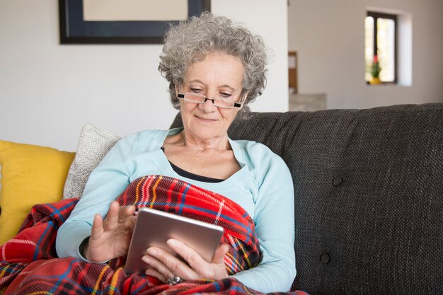 Neugierige ältere Frau, die auf Sofa sitzt und Gerät verwendet