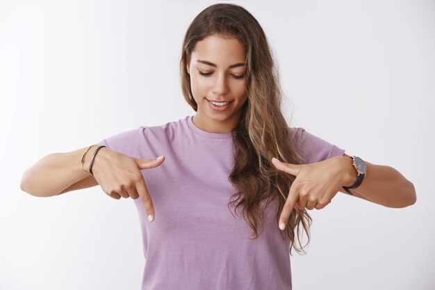 Neugierige attraktive junge gebräunte Frau mit lila T-Shirt, die mit den Zeigefingern nach unten zeigt, amüsiert lächelt, faszinierende tolle Promo nach unten findet und Ihnen gute Promo zeigt