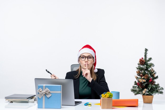 Neugierige blonde Frau mit einem Weihnachtsmannhut, der an einem Tisch mit einem Weihnachtsbaum und einem Geschenk auf ihm auf weißem Hintergrund sitzt