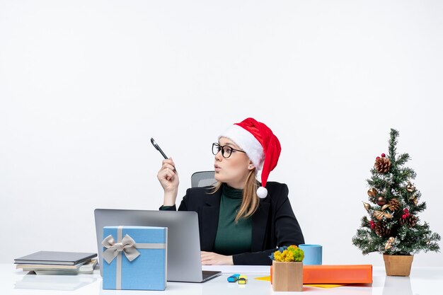 Neugierige blonde Frau mit einem Weihnachtsmannhut, der an einem Tisch mit einem Weihnachtsbaum und einem Geschenk darauf sitzt und etwas auf weißem Hintergrund betrachtet