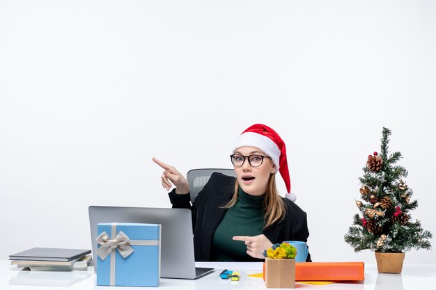 Neugierige junge Frau mit Weihnachtsmannhut, die an einem Tisch mit einem Weihnachtsbaum und einem Geschenk darauf sitzt und oben auf weißem Hintergrund zeigt