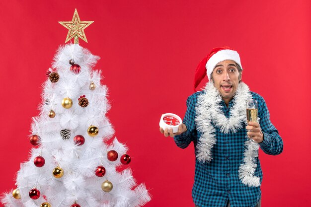 Neugieriger junger Mann mit Weihnachtsmannhut, der ein Glas Wein hebt und Uhr hält, die nahe Weihnachtsbaum steht