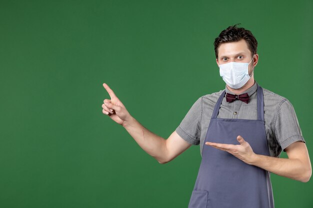 neugieriger Kellner in Uniform mit medizinischer Maske und nach oben auf der rechten Seite auf grünem Hintergrund