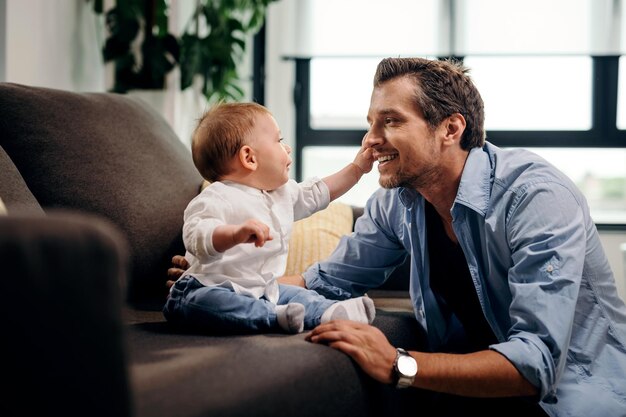 Neugieriger kleiner Sohn, der das Gesicht des Vaters berührt, während er auf dem Sofa im Wohnzimmer sitzt