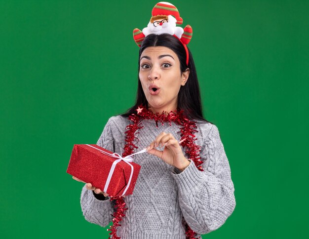 Neugieriges junges kaukasisches Mädchen, das Weihnachtsmann-Stirnband und Lametta-Girlande um den Hals hält, der Geschenkpaket hält, das Kamera-Greifband lokalisiert auf grünem Hintergrund betrachtet