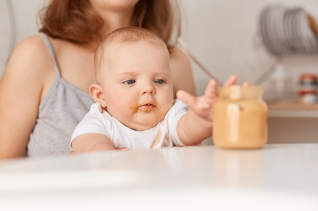 Neugieriges Kind, das den Arm ausstreckt, um das Glas zu füttern, die gesichtslose Mutter füttert ihre kleine Tochter mit Gemüsepüree, sitzt zu Hause am Tisch und füttert.