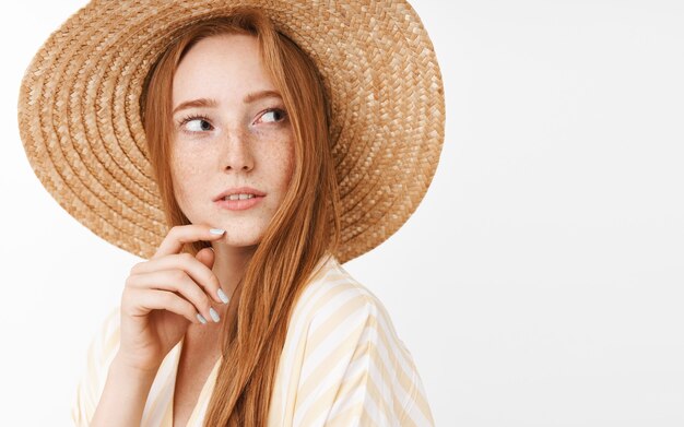 Neugieriges, nachdenkliches, charmantes Ingwer-Mädchen mit niedlichen Sommersprossen im trendigen Sommer-Strohhut, das sich nach rechts dreht und mit Interesse und fasziniertem Blick das Kinn berührt, während es denkt und beobachtet