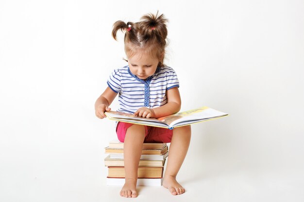 Neugieriges weibliches Kind mit zwei lustigen Pferdeschwänzen, sitzt auf Stapel Bücher, liest interessante Märchen, sieht bunte Bilder mit großem Interesse an, lernt isoliert auf weißem Studio zu lesen.