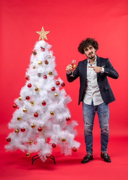 Neujahrsfeier mit bärtigem jungen Mann, der Wein zeigt und nahe verziertem weißem Weihnachtsbaum steht