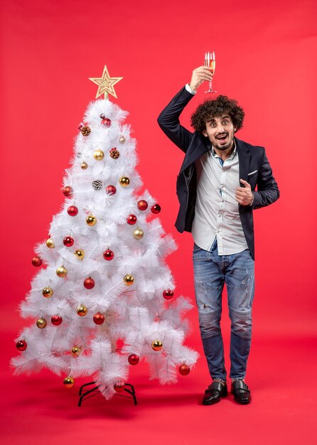 Neujahrsfeier mit jungem Mann, der ein Glas Wein auf seinem Kopf nahe verziertem weißem Weihnachtsbaum hält