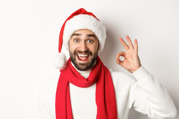 Neujahrsparty und Winterferienkonzept. Nahaufnahme eines glücklichen, attraktiven Mannes in Weihnachtsmütze, der ein gutes Zeichen zeigt und Weihnachten feiert, weißer Hintergrund