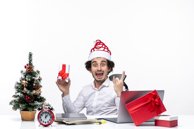 Neujahrsstimmung mit aufgeregtem emotionalem jungem Geschäftsmann mit Weihnachtsmannhut, der im Büro sitzt und sein Geschenk hält, das für Kamera auf weißem Hintergrund aufwirft