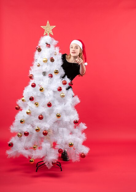 Neujahrsstimmung mit der jungen Frau im schwarzen Kleid und im Weihnachtsmannhut, die hinter weißem Weihnachtsbaum stehen