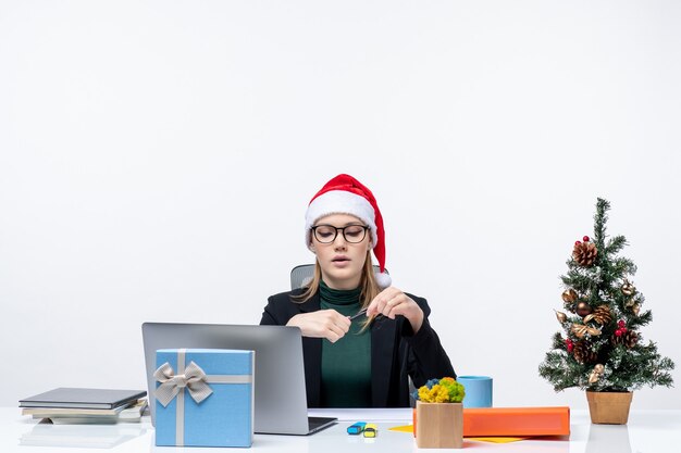 Neujahrsstimmung mit ernsthafter blonder Frau mit einem Weihnachtsmannhut, der an einem Tisch mit einem Weihnachtsbaum und einem Geschenk auf ihm auf weißem Hintergrund sitzt