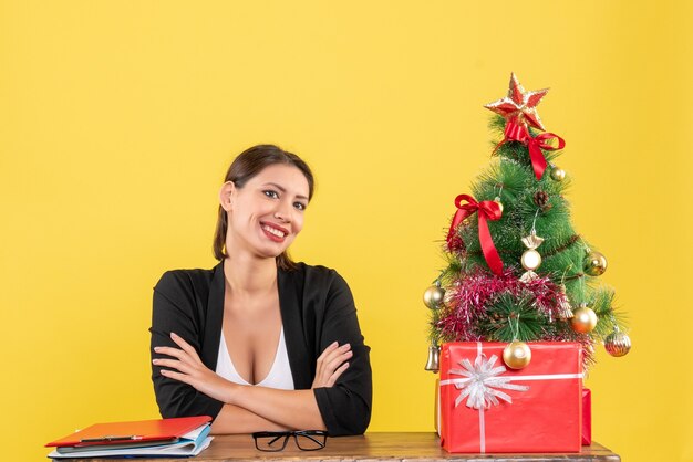 Neujahrsstimmung mit glücklicher junger Frau im Anzug mit geschmücktem Weihnachtsbaum im Büro auf Gelb