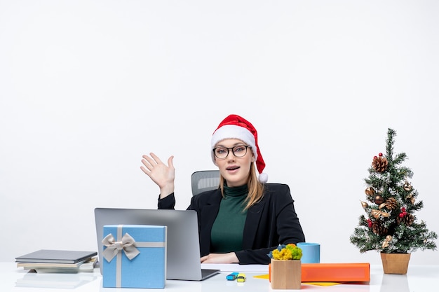 Neujahrsstimmung mit junger attraktiver Frau mit einem Weihnachtsmannhut, der an einem Tisch mit einem Weihnachtsbaum und einem Geschenk darauf sitzt, das über etwas im Büro spricht