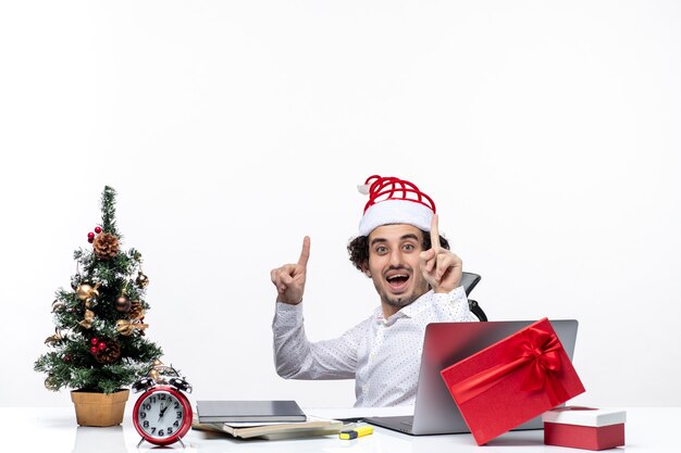 Neujahrsstimmung mit lächelndem aufgeregtem jungem Geschäftsmann, der im Büro sitzt und oben auf weißem Hintergrund zeigt