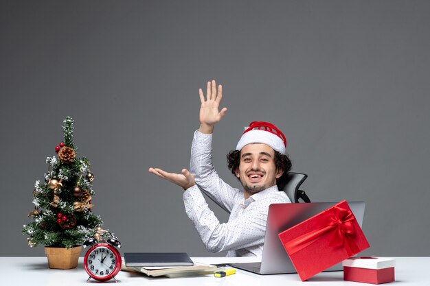 Neujahrsstimmung mit lächelndem positivem jungen Geschäftsmann mit Weihnachtsmannhut, der im Büro sitzt und allein am Projekt auf dunklem Hintergrund arbeitet