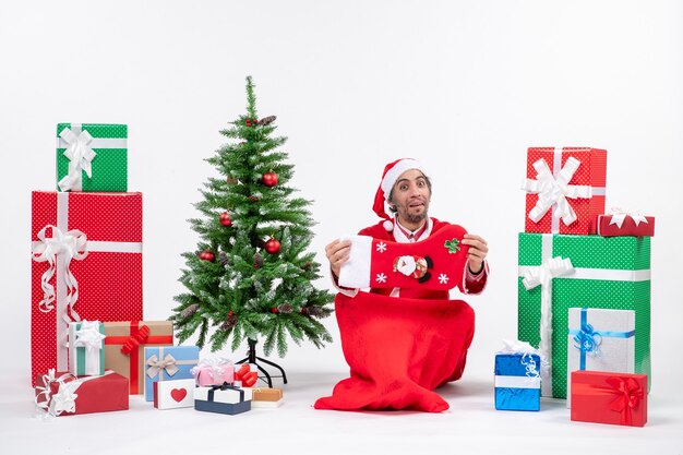 Neujahrsstimmung mit lustigem positivem Weihnachtsmann, der auf dem Boden sitzt und Weihnachtssocke nahe Geschenken und geschmücktem Weihnachtsbaum auf weißem Hintergrund hält