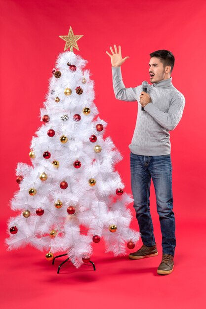 Neujahrsstimmung mit schockiertem positivem Kerl gekleidet in Jeans, die nahe geschmücktem Weihnachtsbaum stehen