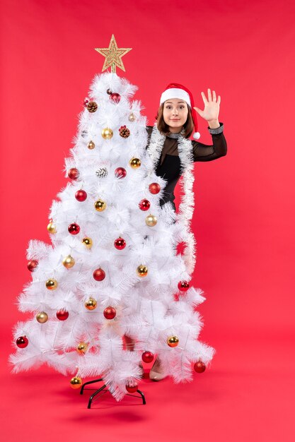 Neujahrsstimmung mit schönem Mädchen in einem schwarzen Kleid mit Weihnachtsmannhut, der sich hinter Weihnachtsbaum versteckt