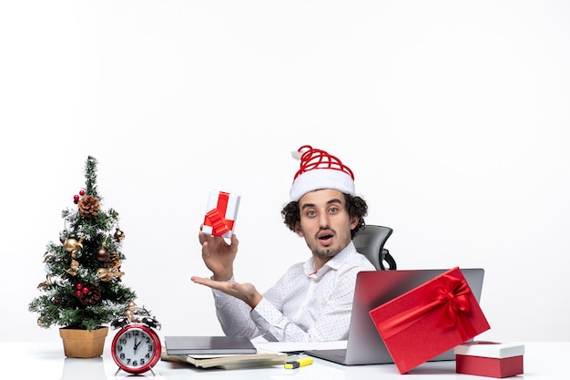 Neujahrsstimmung mit überraschtem jungen Geschäftsmann mit Weihnachtsmannhut, der im Büro sitzt und sein Geschenk hält, das für Kamera auf weißem Hintergrund aufwirft