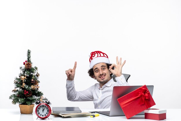 Neujahrsstimmung mit überraschtem lächelndem aufgeregtem aufgeregtem jungen Geschäftsmann, der im Büro sitzt und perfekte Geste auf weißem Hintergrund macht