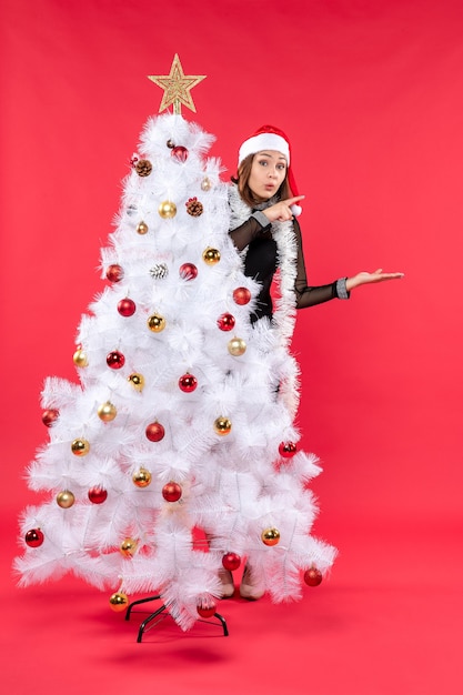 Neujahrsstimmung mit überraschtem schönem Mädchen in einem schwarzen Kleid mit Weihnachtsmannhut, der sich hinter Weihnachtsbaum versteckt