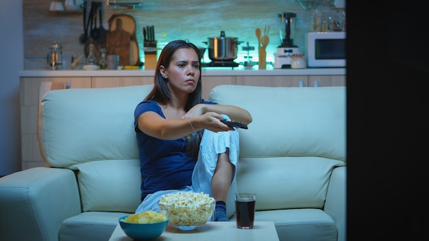Nevous Dame wechselt die Kanäle auf einer gemütlichen Couch. Gelangweilte, wütende allein zu Hause spät in der Nacht Frau entspannt vor dem Fernseher liegend auf einem bequemen Sofa mit Fernbedienung und sucht nach einem Comedy-Film.