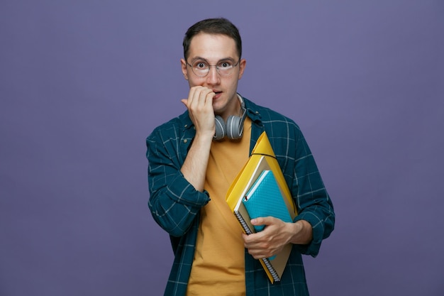 Ängstlich junger männlicher Student, der eine Brille mit Kopfhörern um den Hals trägt und einen Notizbuch-Notizblock-Ordner hält, der in die Kamera schaut, beißende Finger isoliert auf violettem Hintergrund