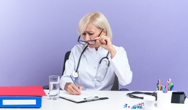 Ängstliche erwachsene Ärztin in optischer Brille, die ein medizinisches Gewand mit Stethoskop trägt und am Schreibtisch mit Bürowerkzeugen sitzt und auf die Zwischenablage schaut, die auf lila Wand mit Kopienraum isoliert ist