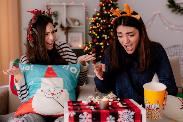 Ängstliche hübsche junge Mädchen mit Stechpalmenkranz und Rentierstirnband schauen auf fallendes Popcorn, das auf Sesseln sitzt und die Weihnachtszeit zu Hause genießt