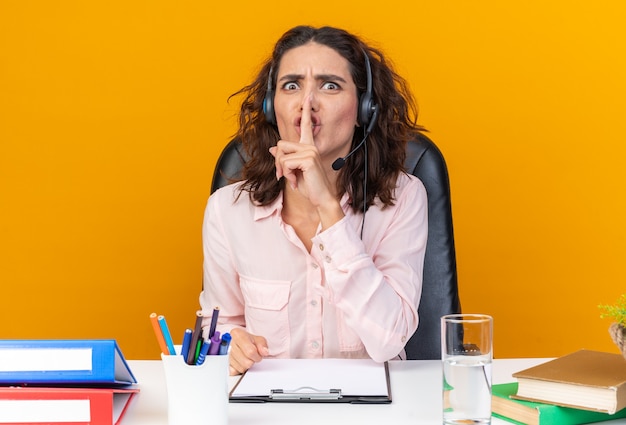 Ängstliche hübsche kaukasische Callcenter-Betreiberin auf Kopfhörern, die am Schreibtisch mit Bürowerkzeugen sitzen, die Stillegeste einzeln auf oranger Wand tun?