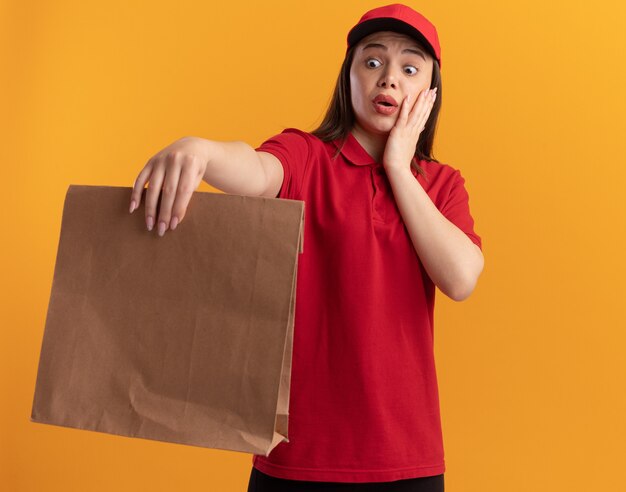 Ängstliche hübsche Lieferfrau in Uniform legt die Hand auf das Gesicht, das Papierpaket hält und betrachtet