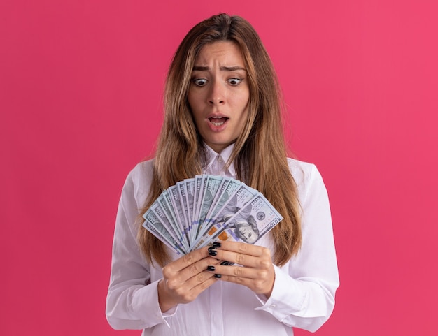 Ängstliche junge hübsche kaukasische Mädchen hält und betrachtet Geld isoliert auf rosa Wand mit Kopierraum