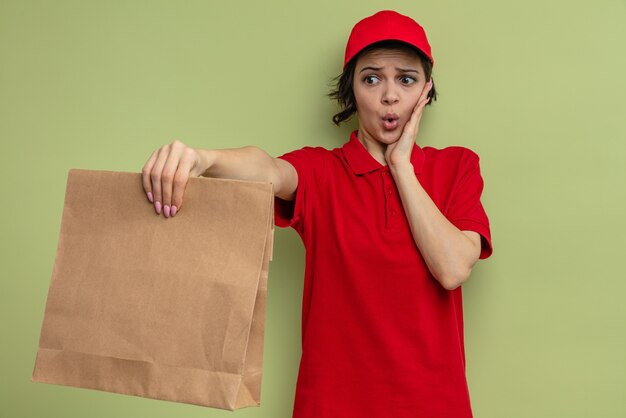 Ängstliche junge hübsche Lieferfrau, die sich die Hand aufs Gesicht legt und Papierverpackungen für Lebensmittel hält