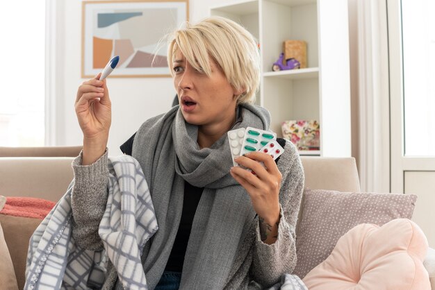 Ängstliche junge kranke slawische Frau mit Schal um den Hals, eingewickelt in Plaid, die Medizinblisterpackungen hält und auf das Thermometer schaut, das auf der Couch im Wohnzimmer sitzt