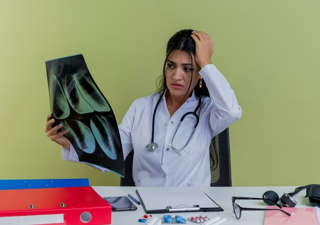 Ängstliche junge Ärztin, die medizinische Robe und Stethoskop trägt, die am Schreibtisch mit medizinischen Werkzeugen sitzen und Röntgenaufnahme halten und Hand auf Kopf isoliert halten