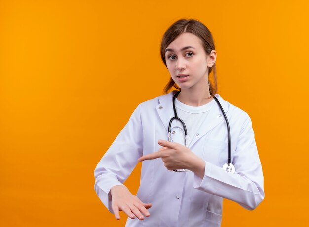 Ängstliche junge Ärztin, die medizinische Robe und Stethoskop trägt, zeigt auf ihre Hand auf lokalisiertem orange Raum mit Kopienraum