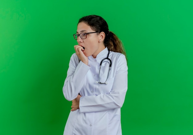 Ängstliche junge Ärztin, die medizinische Robe und Stethoskop und Brille trägt, die Seitenbeißfinger lokalisiert auf grüner Wand mit Kopienraum betrachten