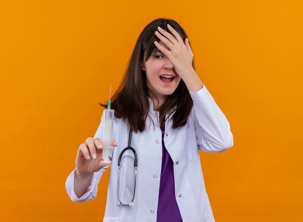 Ängstliche junge Ärztin im medizinischen Gewand mit Stethoskop hält Spritze und hält Kopf auf lokalisiertem orangefarbenem Hintergrund mit Kopienraum