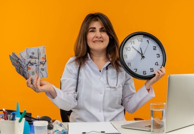 Ängstliche Ärztin mittleren Alters, die medizinische Robe und Stethoskop trägt, sitzt am Schreibtisch mit medizinischen Werkzeug-Zwischenablage und Laptop, die Uhr und Geldbeißlippe isoliert halten