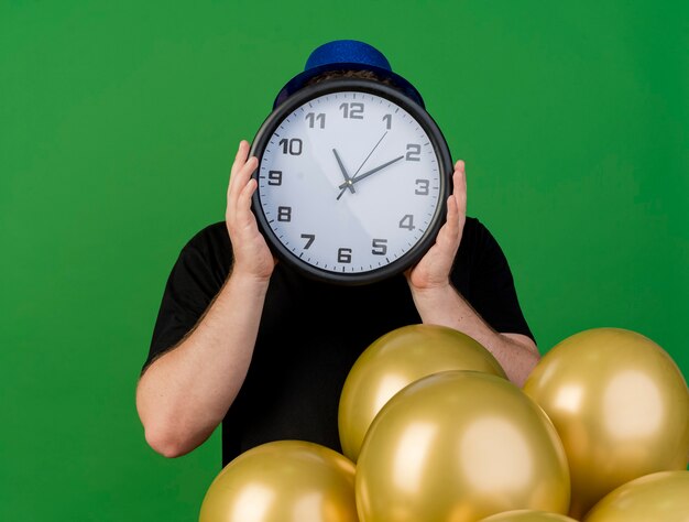 Ängstlicher erwachsener Mann in der optischen Brille, die blauen Parteihut trägt, hält Uhr vor Gesicht und steht mit Heliumballons isoliert auf grüner Wand