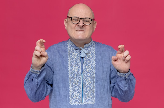 Ängstlicher erwachsener slawischer Mann im blauen Hemd mit optischer Brille, die Finger kreuzt und