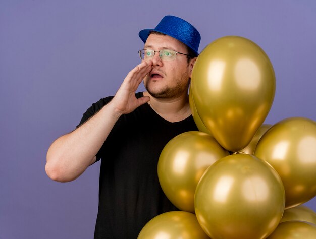 Ängstlicher erwachsener slawischer Mann in optischer Brille mit blauem Partyhut hält die Hand nah am Mund und hält Heliumballons