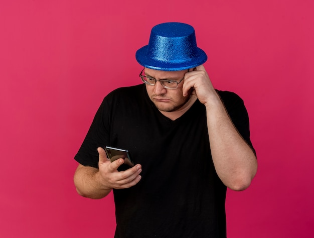 Ängstlicher erwachsener slawischer Mann in optischer Brille mit blauem Partyhut hält und schaut auf das Telefon