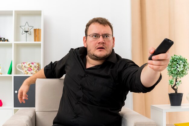 Ängstlicher erwachsener slawischer Mann mit optischer Brille sitzt auf einem Sessel, der eine TV-Fernbedienung hält