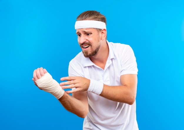 Ängstlicher junger, gutaussehender, sportlicher Mann mit Stirnband und Armbändern, der sein verletztes Handgelenk betrachtet, das mit Verband isoliert auf blauer Wand gewickelt ist
