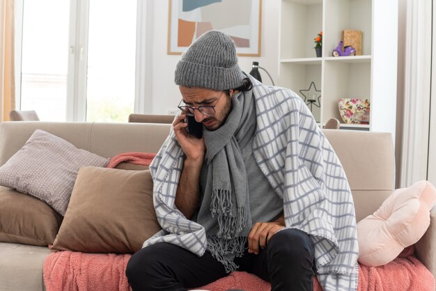 Ängstlicher junger kranker Mann in optischer Brille, eingewickelt in Plaid mit Schal um den Hals, der eine Wintermütze trägt und am Telefon spricht, sitzt auf der Couch im Wohnzimmer